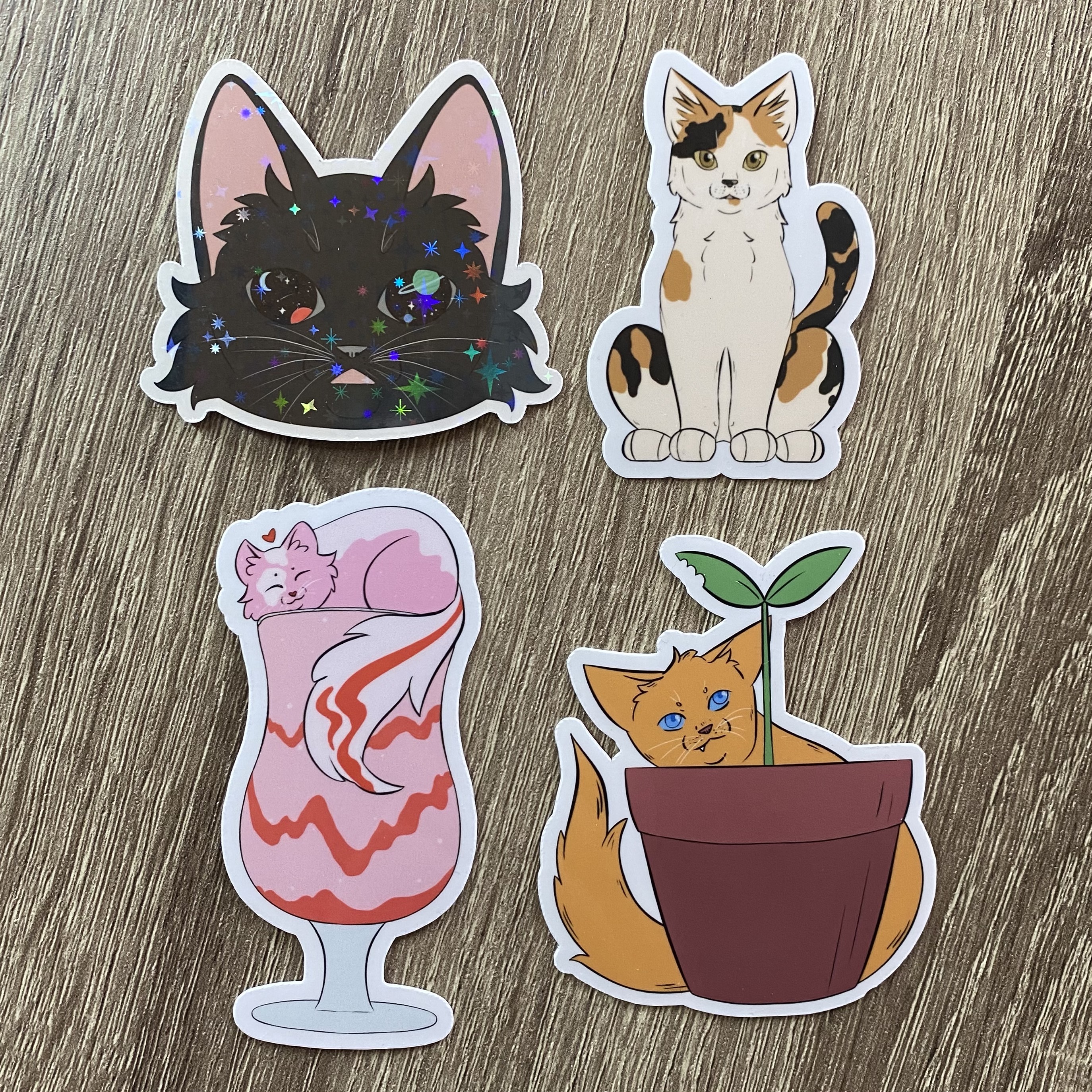 Assorted cat stickers, the first is a sparkly black cat with space eyes, the second is a glossy calico cat, the third is a glossy cat on a milkshake and the fourth is a glossy cat with a potted plant.